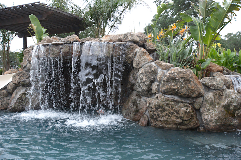 A waterfall in a pool in a backyard with tropical landscaping